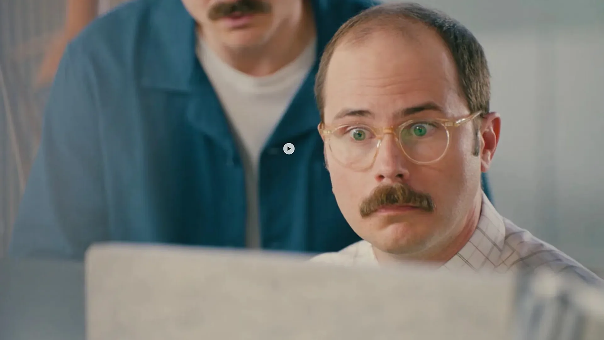 A wide eyed man with a mustache and round clear glasses stares at a laptop screen, a blurred man in a teal jacket behind him. A small circular play button sits at the center.