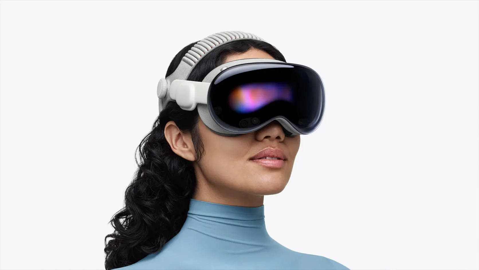 A woman with long dark curly hair wears a white and grey mixed reality headset, its curved dark visor glowing pink and blue, over a pale blue turtleneck on a white background.