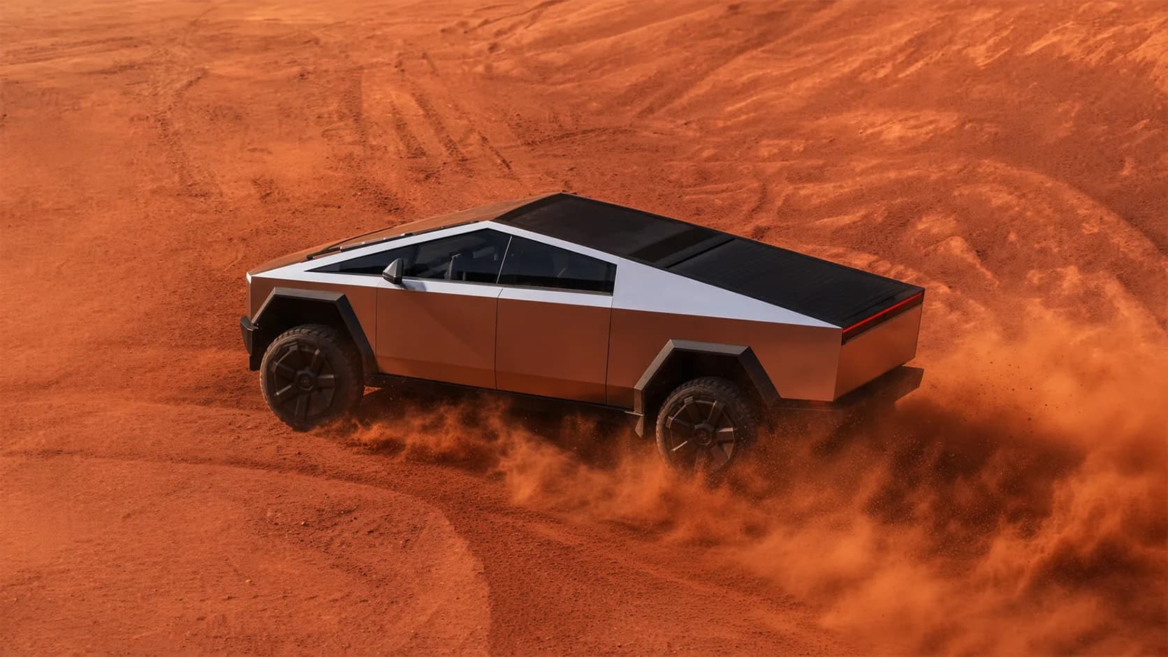 An angular stainless steel pickup truck with a sharp wedge profile drives across rust red desert sand, seen from above, kicking up dust behind its rear wheels beside curving tire tracks.