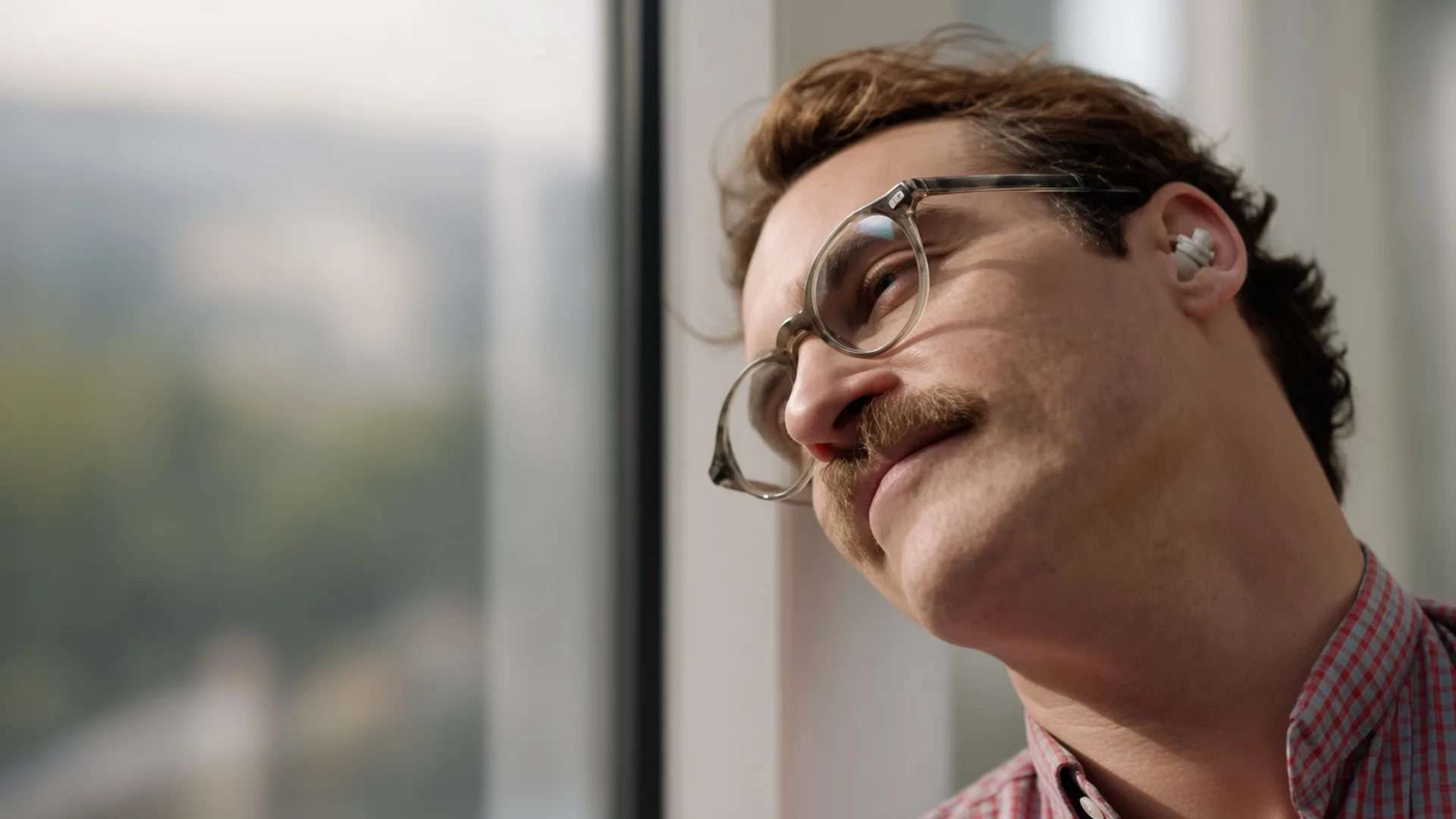 Close crop of a man with a thick moustache, round clear framed glasses and a small white earpiece, smiling faintly as he gazes past a blurred window in a red and blue checked shirt.