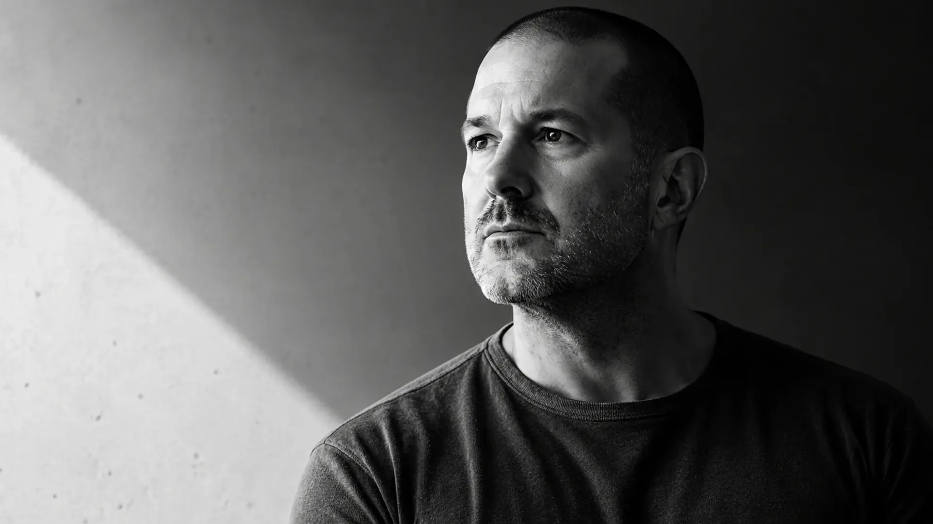 A black and white portrait of a person with a shaved head and stubble in a dark t-shirt, beside a light-streaked concrete wall.