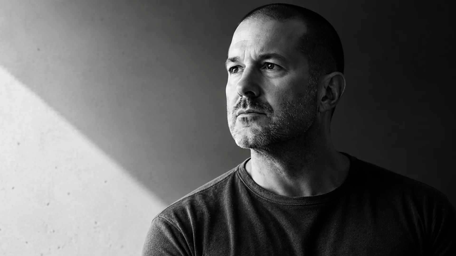 Black and white portrait of a man with a closely shaved head and stubble in a plain dark tee, looking off to the left, against a concrete wall crossed by a diagonal shaft of light.