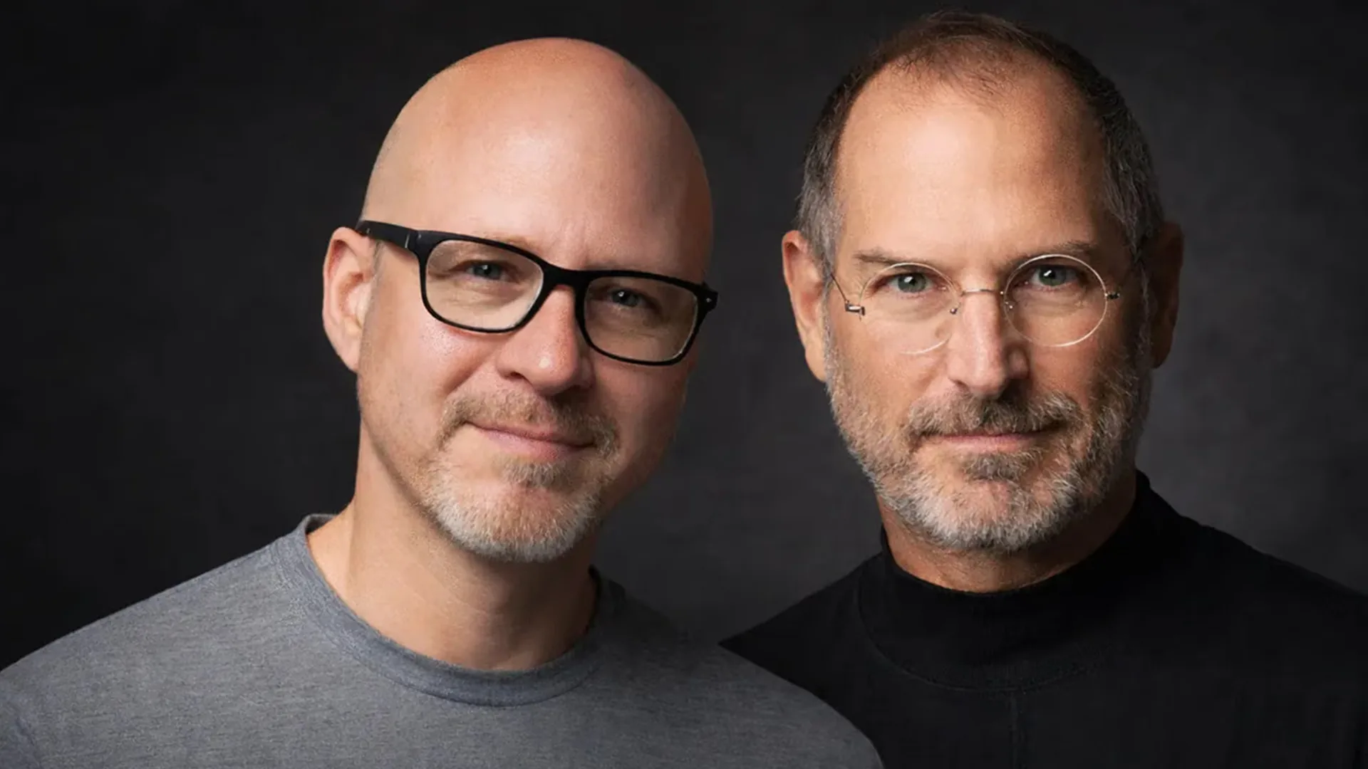 Two men stand shoulder to shoulder against a dark grey studio backdrop, the left one bald in black rectangular glasses and a grey sweatshirt, the right one greying in rimless round glasses and a black turtleneck.