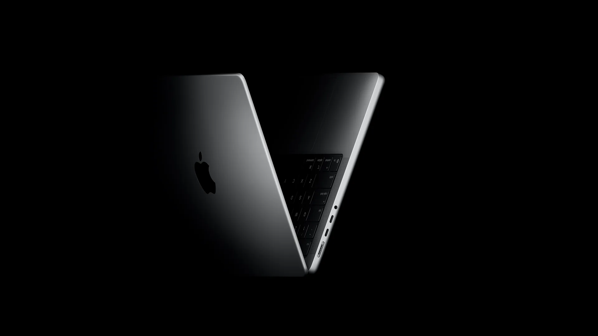 A silver Apple laptop stands half open in near darkness, its lid angled toward the camera with the Apple logo catching a soft highlight and the keyboard and side ports faintly lit.