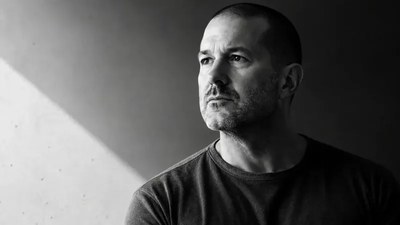 A black and white portrait of a person with a shaved head and stubble in a dark t-shirt, beside a light-streaked concrete wall.