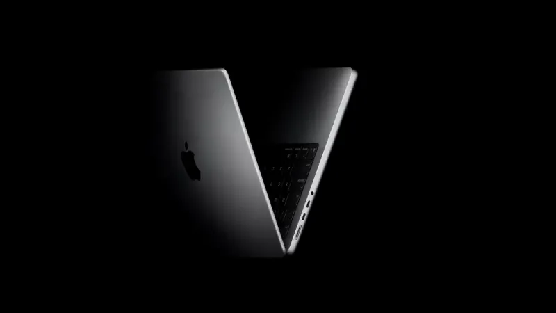 A silver Apple laptop stands half open in near darkness, its lid angled toward the camera with the Apple logo catching a soft highlight and the keyboard and side ports faintly lit.